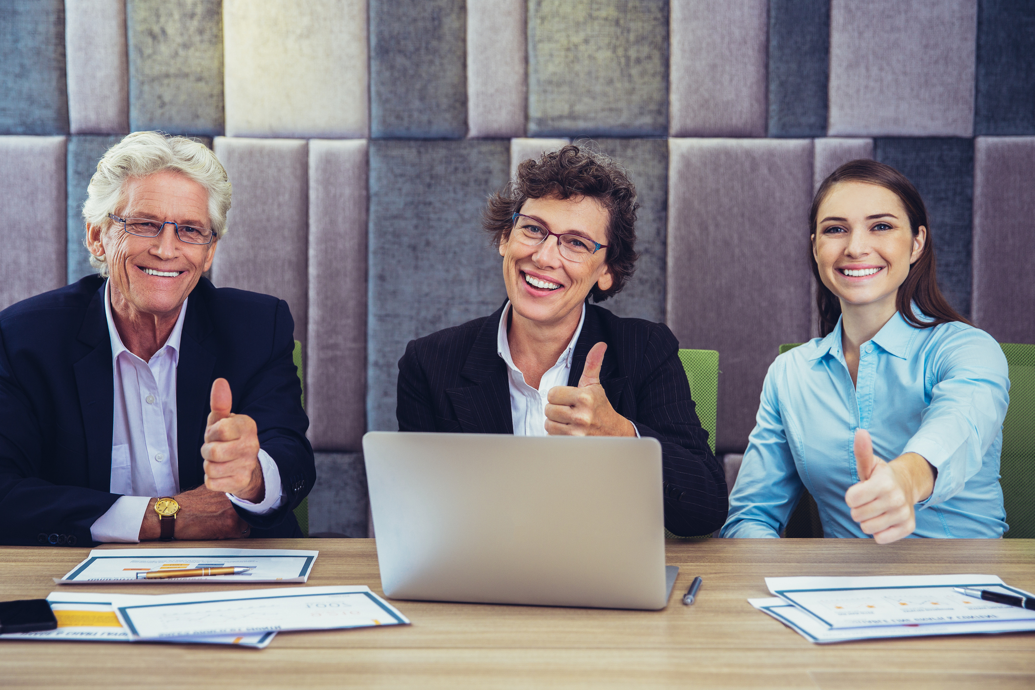 Portrait of positive Caucasian business team, senior woman, young woman and senior man, sitting at table in office, showing thumb up, looking at camera and smiling. Laptop and documents on table