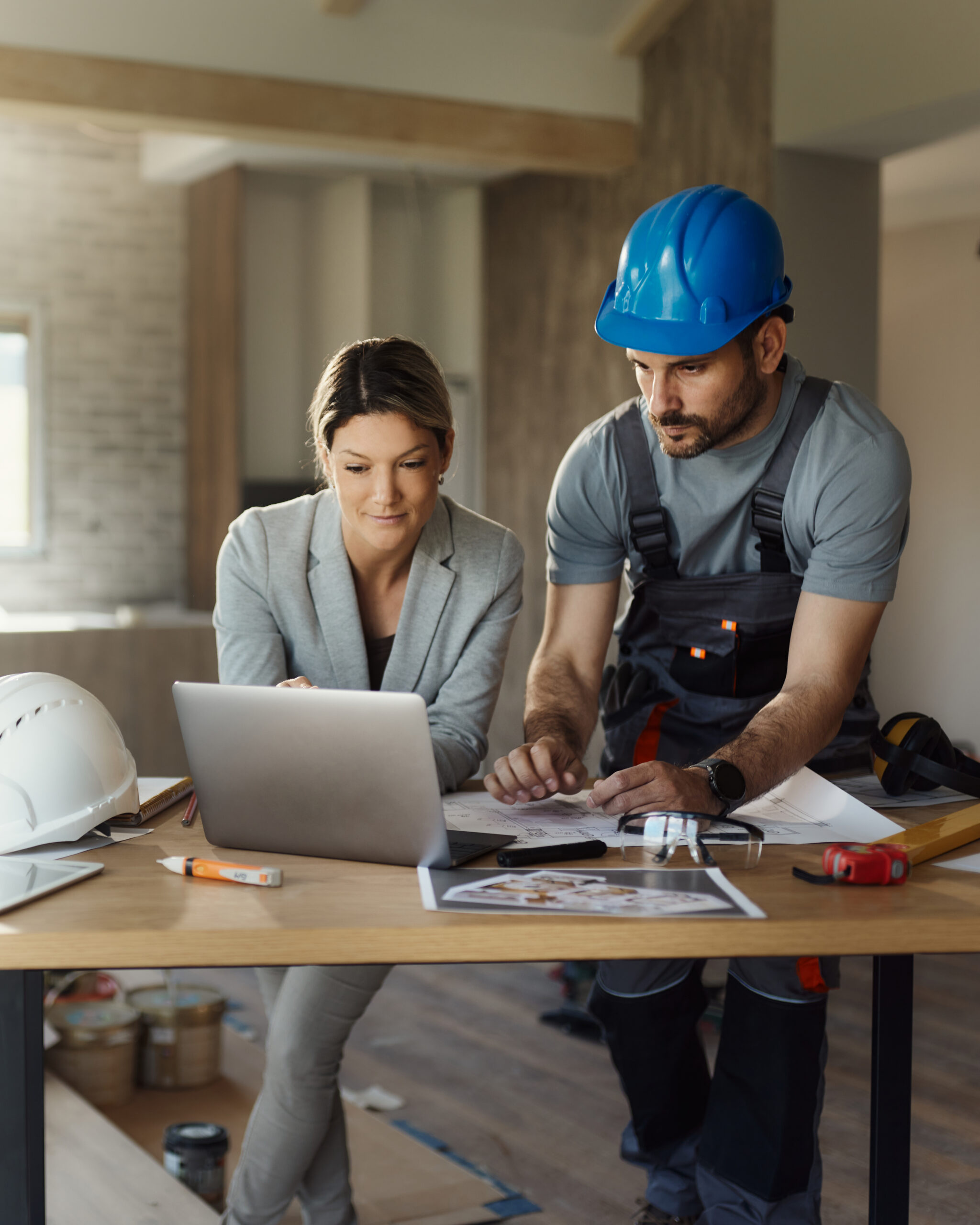 Female interior designer and manual worker cooperating while working on a computer during home renovation process.