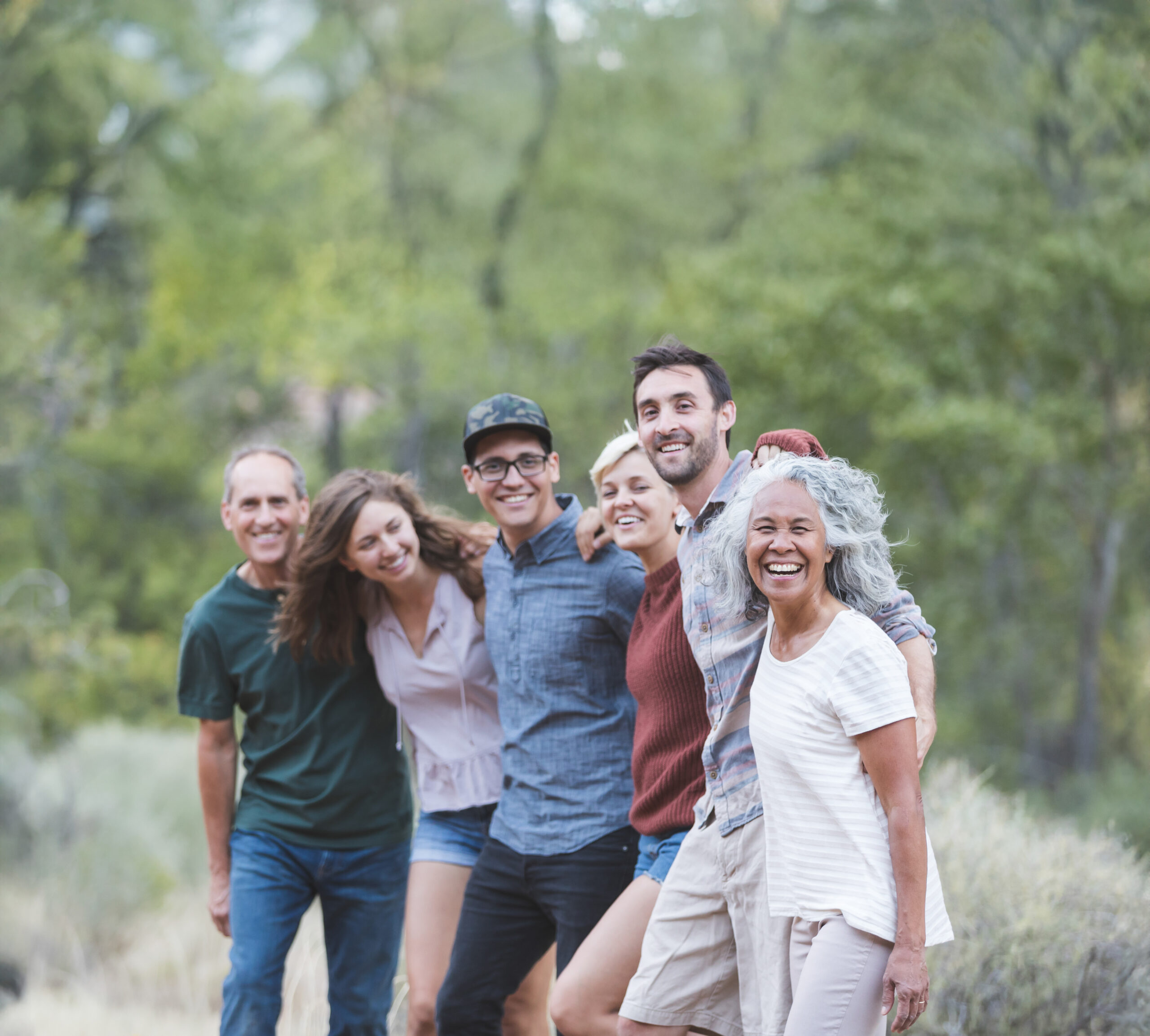 A multiethnic group of friends embrace and smile toward the camera. They're lined up loosely and desert vegetation and sagebrush are in the background.