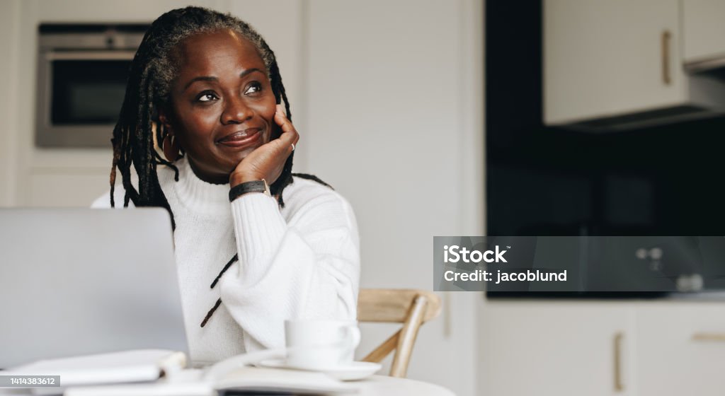 Mature businesswoman looking away thoughtfully while working on a laptop at home. Senior businesswoman doing online freelance work in her home office.
