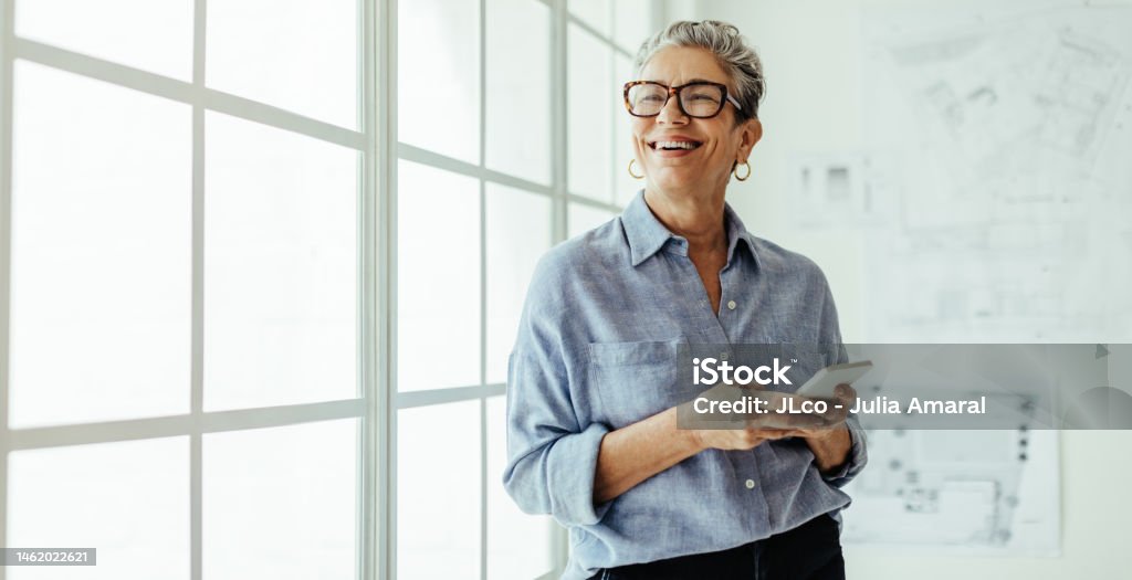 Mature business woman stands in an office, holding a smartphone and looking outside the window with a smile. Female designer using mobile technology to stay connected with her colleagues and clients.