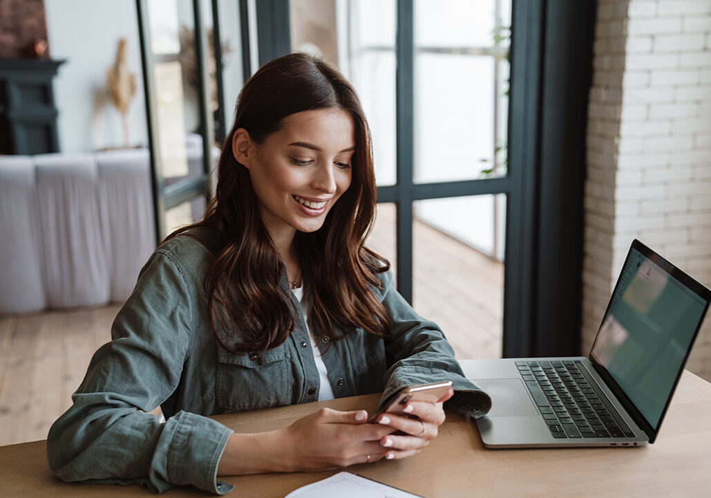 Beautiful smiling woman using cellphone while working with laptop indoors