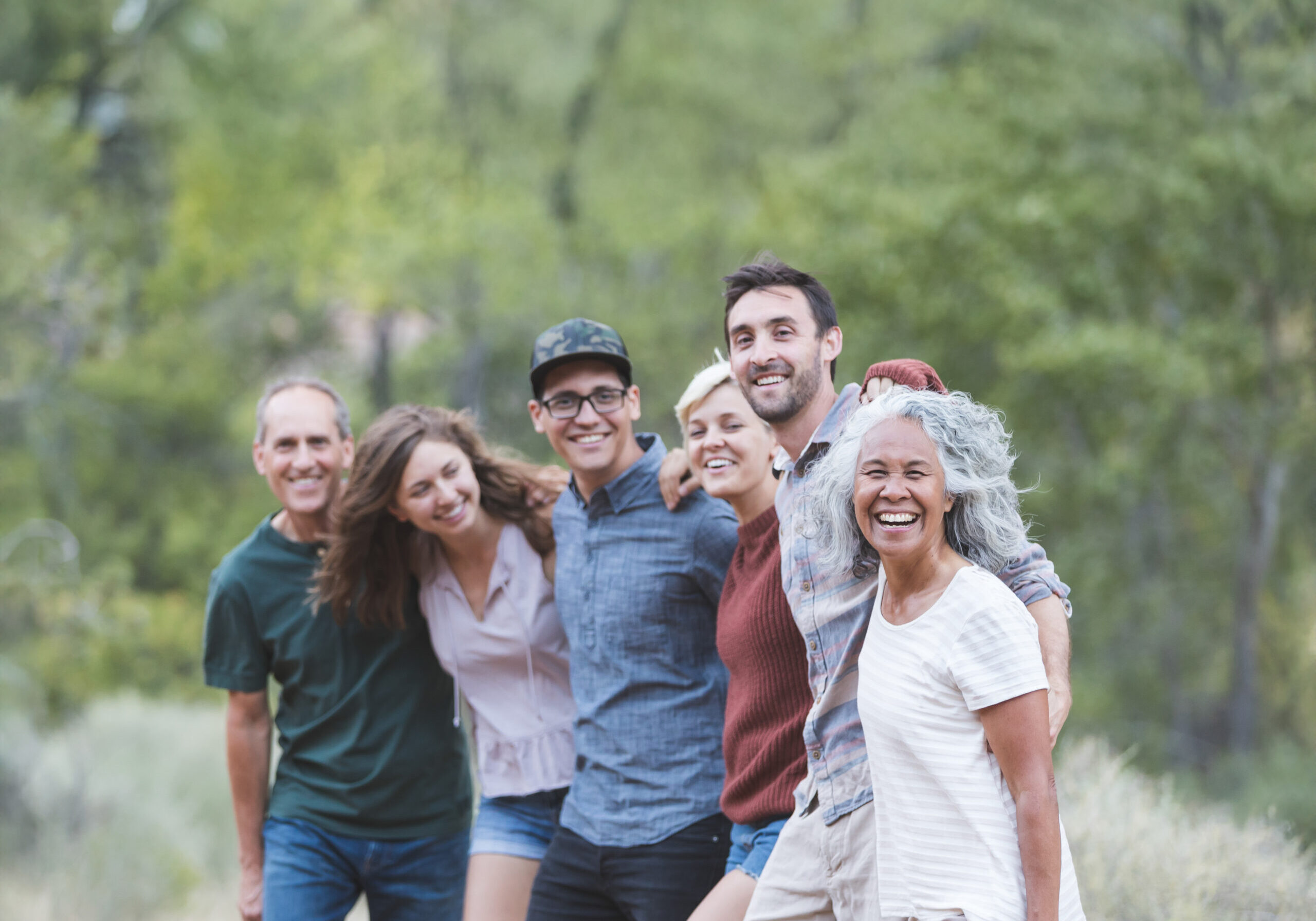 A multiethnic group of friends embrace and smile toward the camera. They're lined up loosely and desert vegetation and sagebrush are in the background.