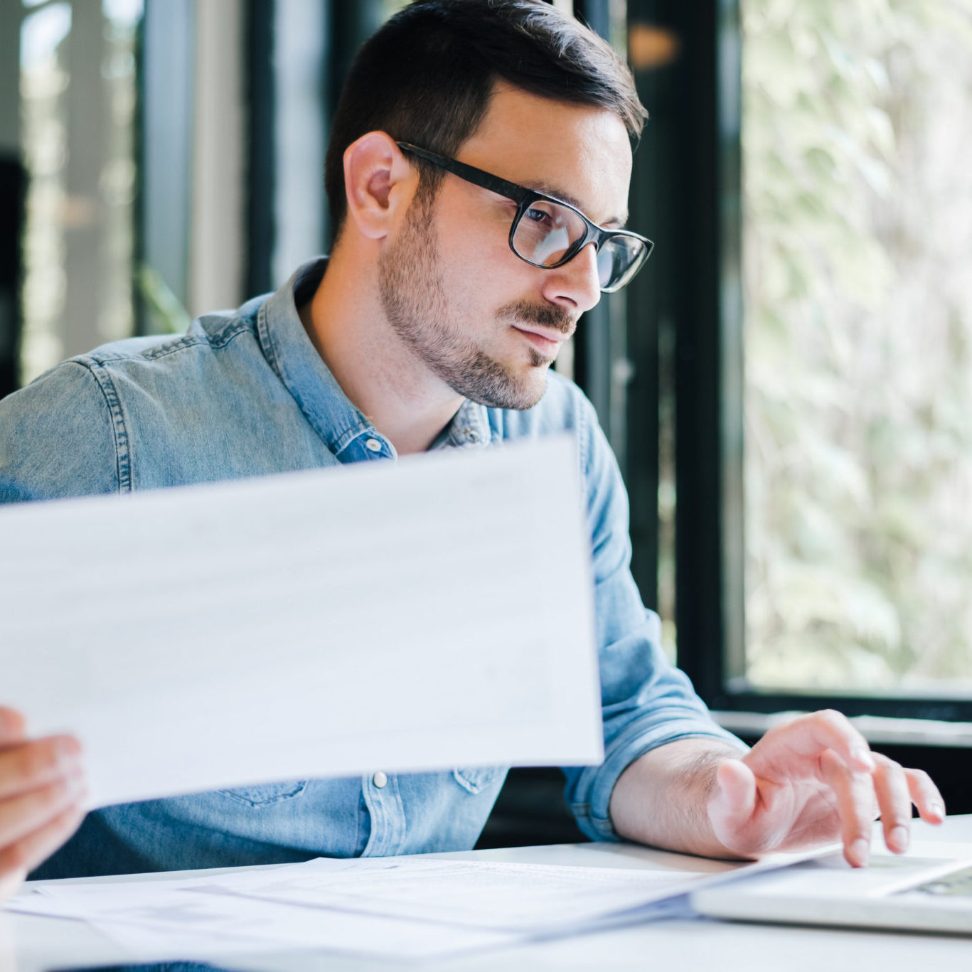 Serious pensive thoughtful focused young casual business accountant bookkeeper in office looking at and working with laptop and income tax return papers and documents